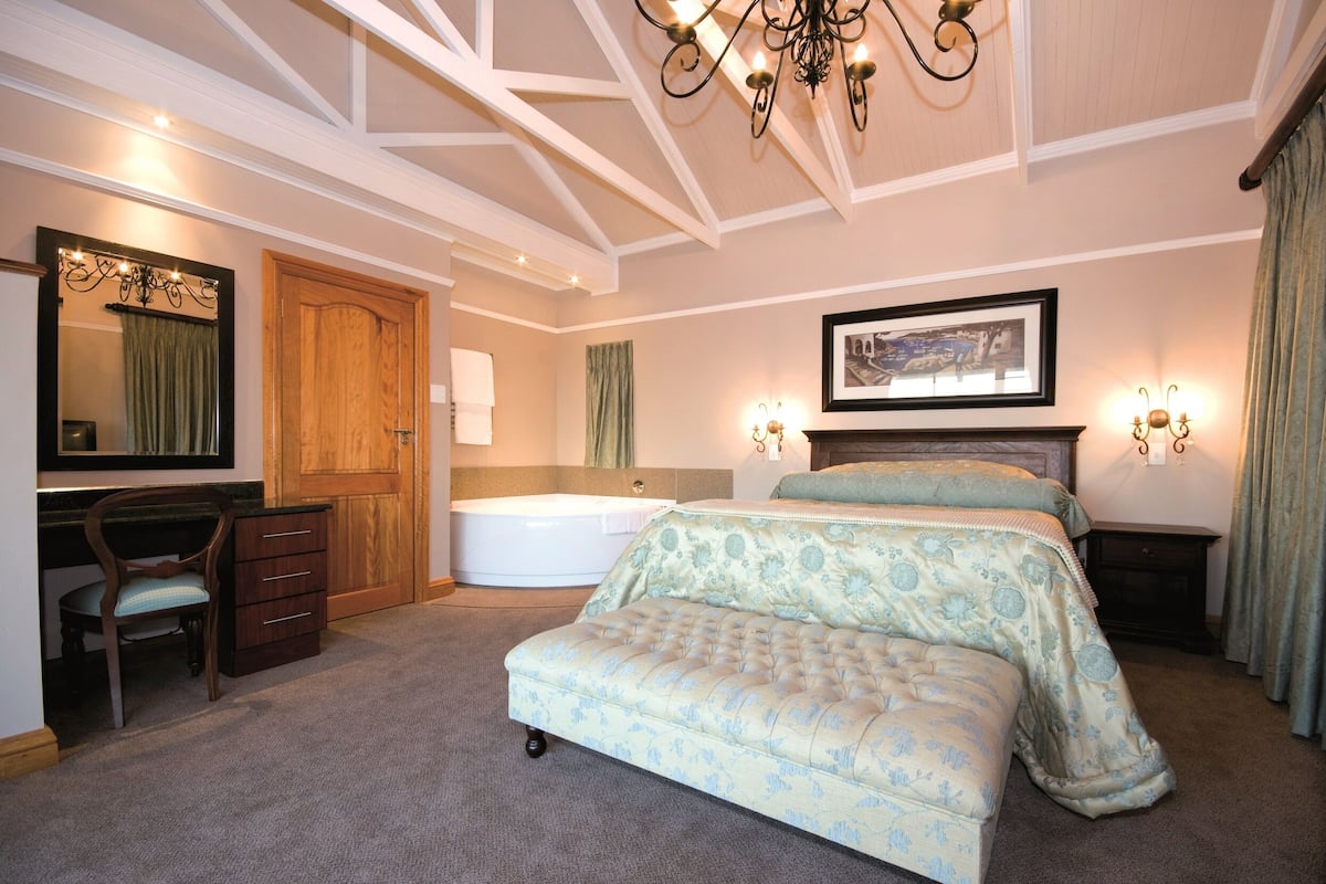 A bedroom with a large bed, a desk with a chair, a wooden wardrobe, a bathtub, and a chandelier.