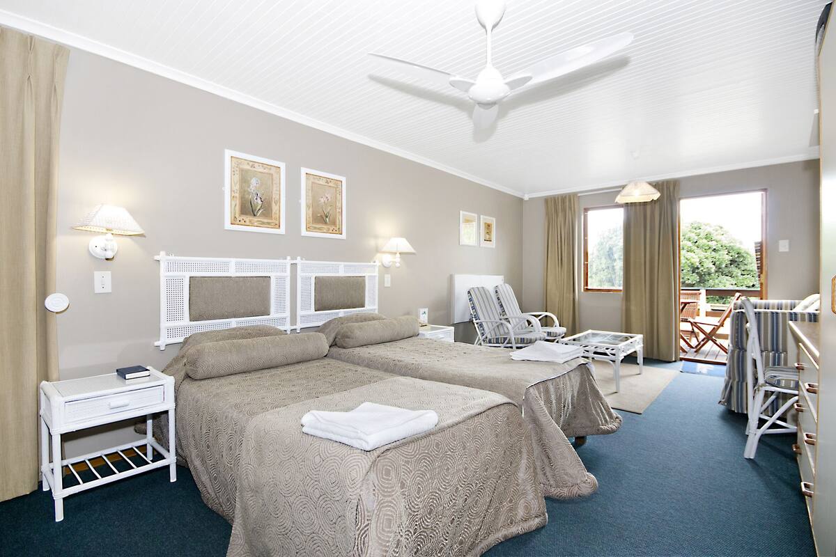 A hotel room with two beds, a ceiling fan, a small table, a chair, and a view of the outdoors.