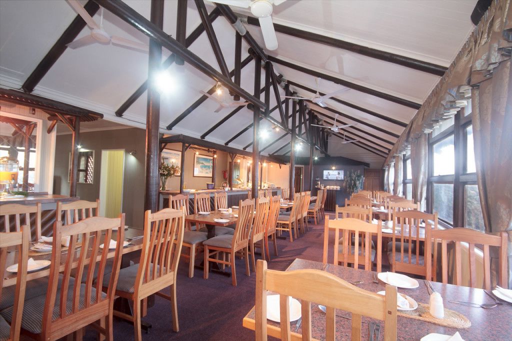 A restaurant with wooden tables and chairs, a high ceiling with exposed beams, and large windows allowing natural light.
