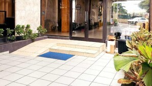 Una entrada moderna a un edificio con puertas de vidrio, un patio con baldosas y un pequeño jardín con plantas.