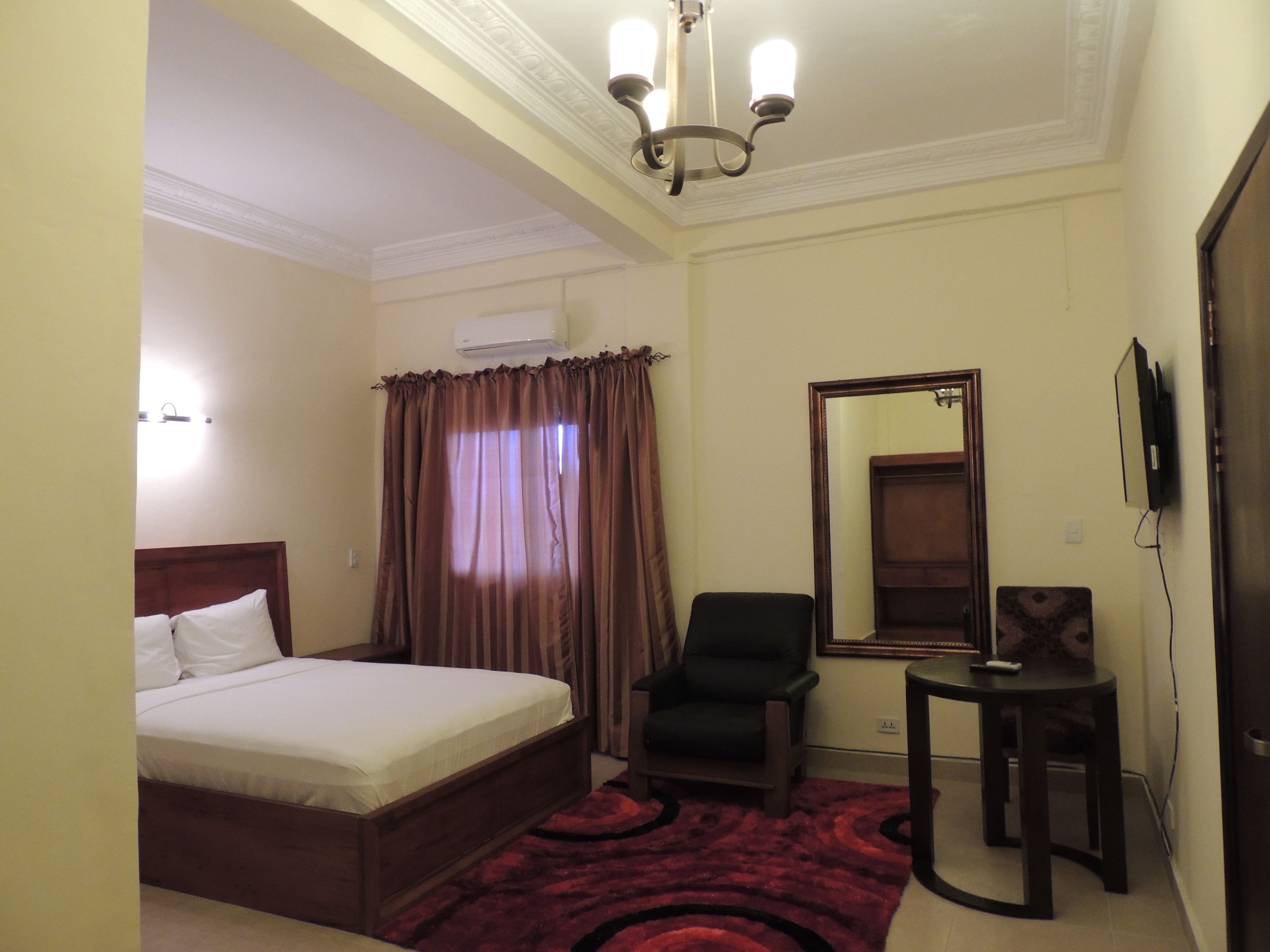 A hotel room with a bed, a chair, a small table, a mirror, and an air conditioning unit.