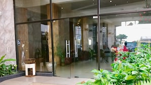 Una entrada moderna de edificio con puerta de cristal, un banco pequeño de madera y exuberantes plantas verdes en primer plano.