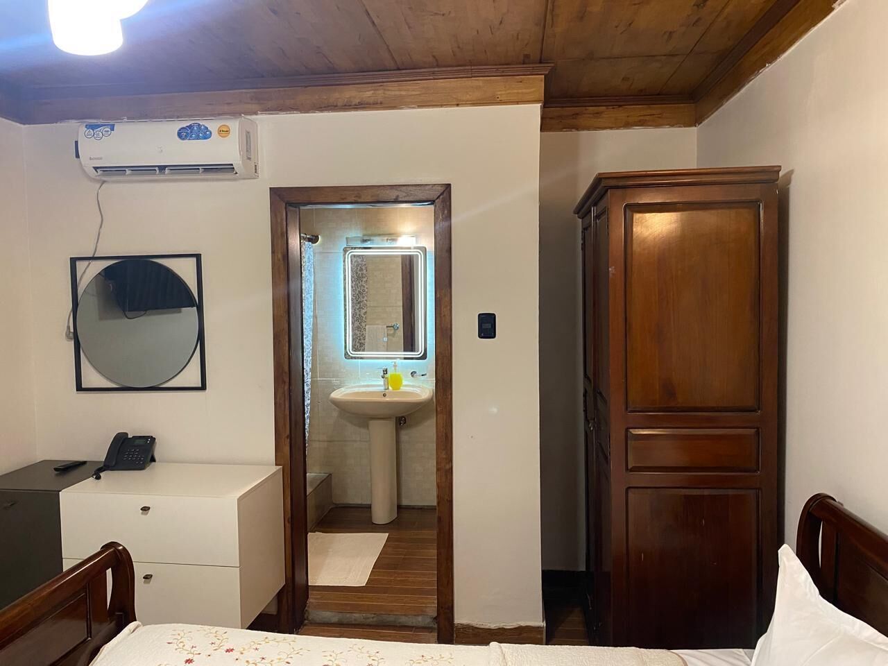 Une chambre à coucher comprenant un lit, une armoire en bois, un bureau avec un téléphone, un miroir et une salle de bain avec un lavabo et un miroir.