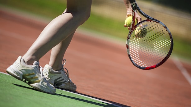 Eine Nahaufnahme der Füße eines Tennisspielers auf einem Sandplatz, der einen Schläger und einen Ball hält.