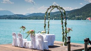 A wooden pier with white chairs and a table, decorated with floral arrangements, overlooking a lake with mountains in the background.