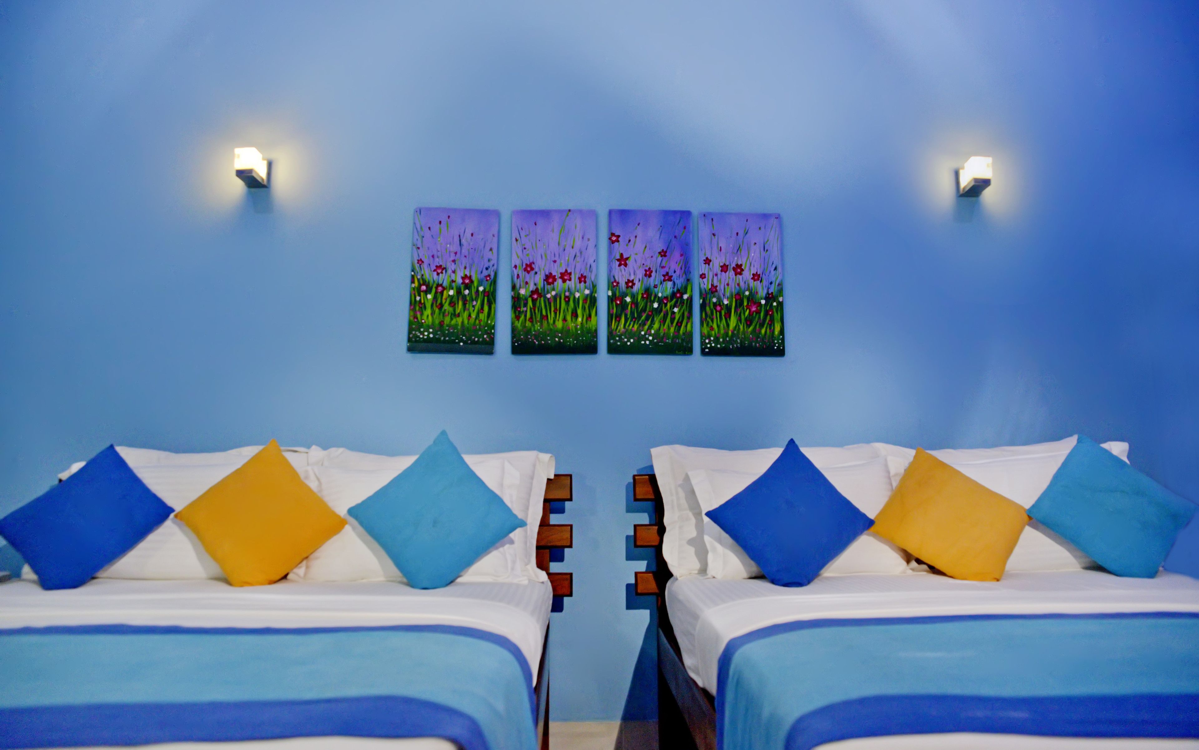 A hotel room with two beds, each with blue and yellow pillows, and a blue wall with three flower paintings.