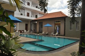 A swimming pool with lounge chairs and umbrellas, surrounded by a building with balconies and a tiled roof. - Peacock Heritage Anuradhapura (Anuradhapura)