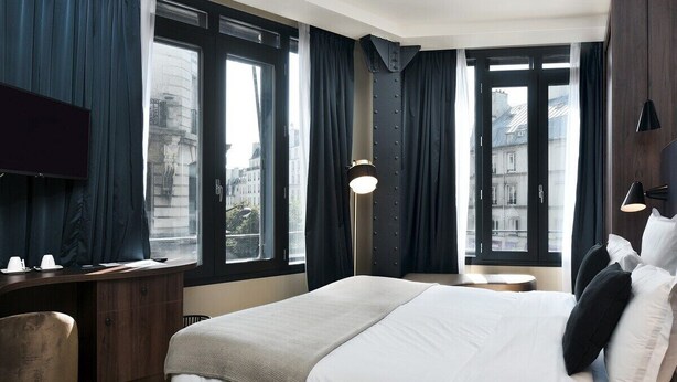 Habitación de hotel con una cama grande, un escritorio, una silla, un televisor y vistas a la ciudad a través de amplios ventanales.