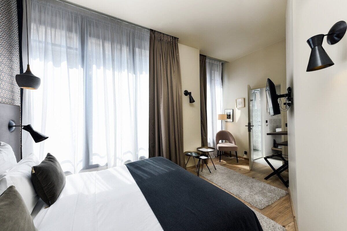 A modern hotel room with a large bed, a desk, and a chair. There is a window with curtains, a lamp, and a small table.