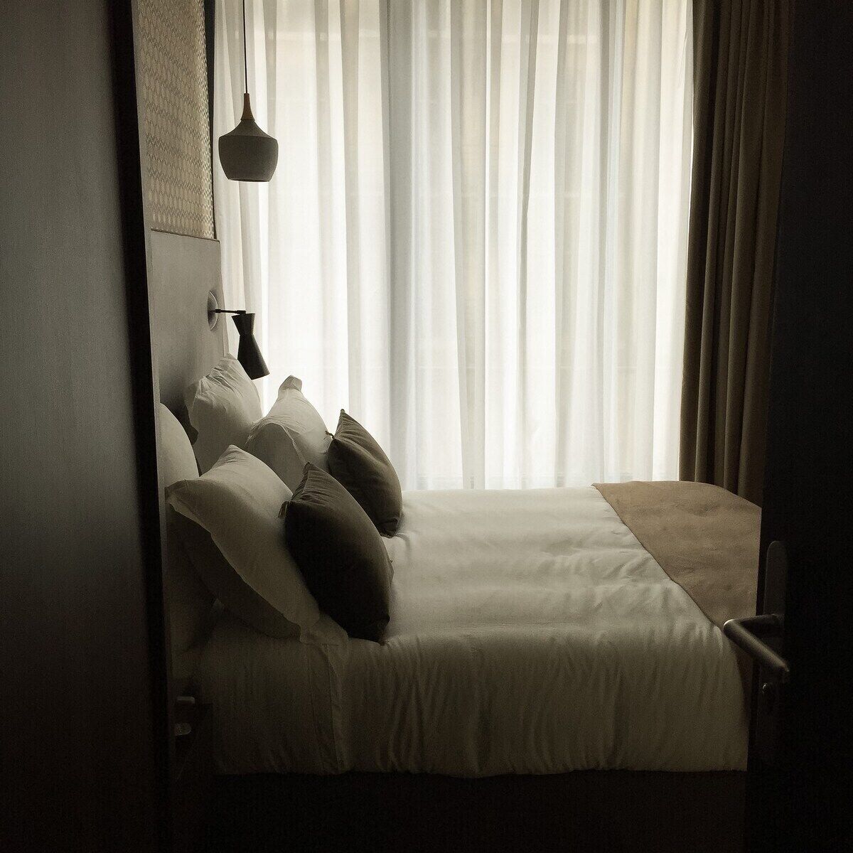 A bedroom with a bed, pillows, a hanging light fixture, and sheer curtains.