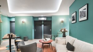 Un moderno vestíbulo de hotel con paredes verde mar, un sofá blanco, un sillón gris y una mesa redonda. Hay dos puertas con las indicaciones “Château” y “Soucier”.