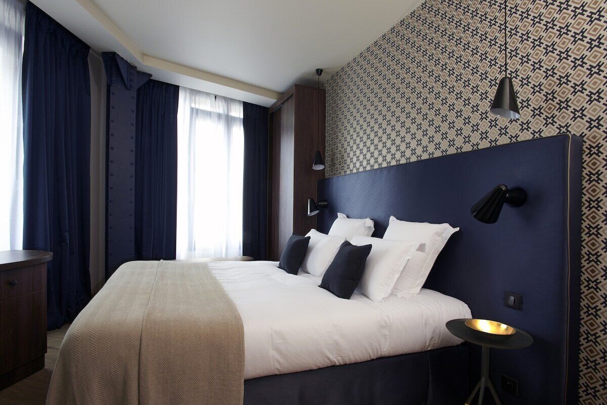 A hotel room with a large bed, dark headboard, patterned wallpaper, and a bedside table with a lamp.
