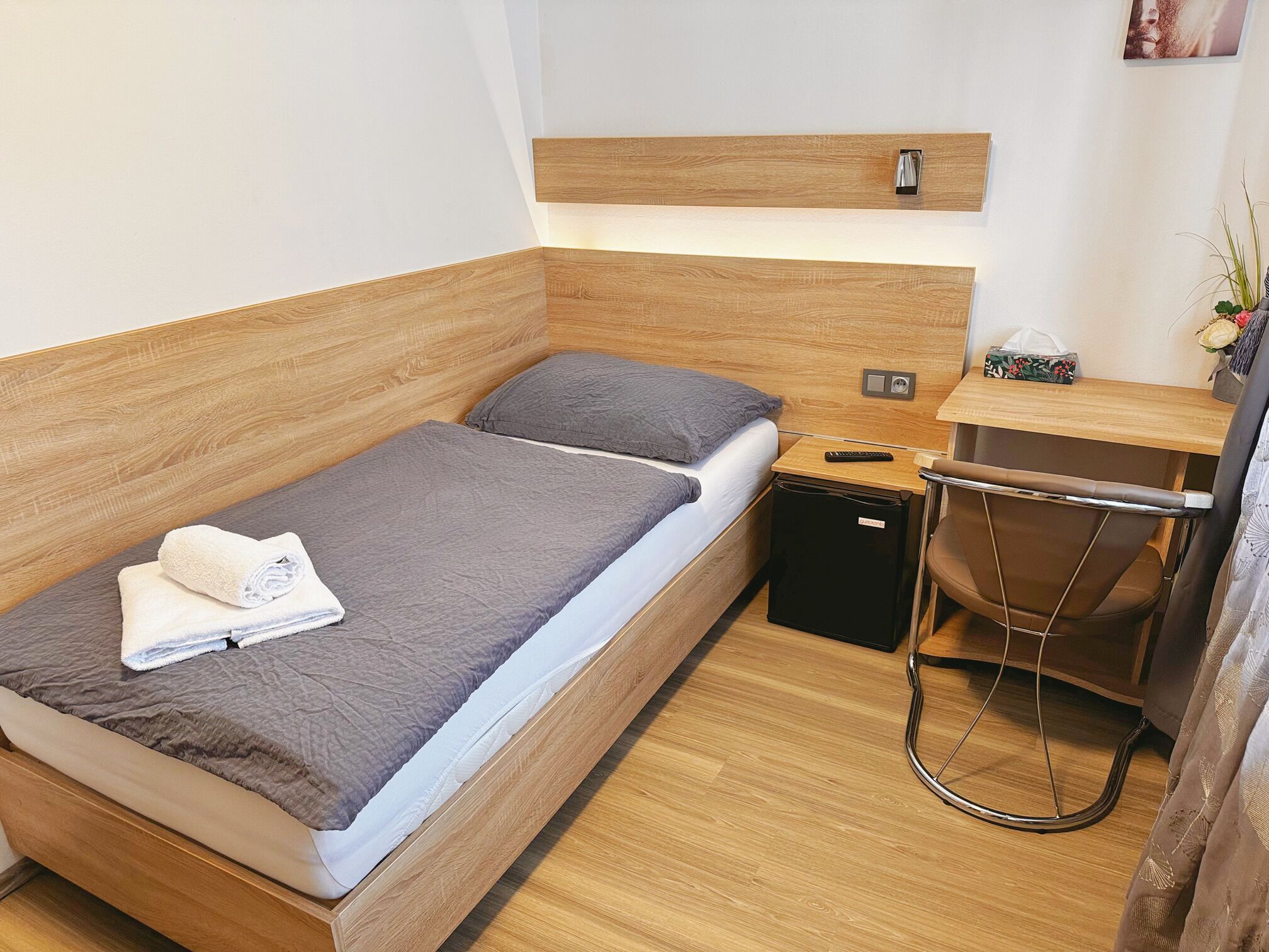 A compact bedroom with a wooden headboard, a single bed with a grey blanket, a small bedside table with a remote control, a compact black storage unit, and a foldable chair.