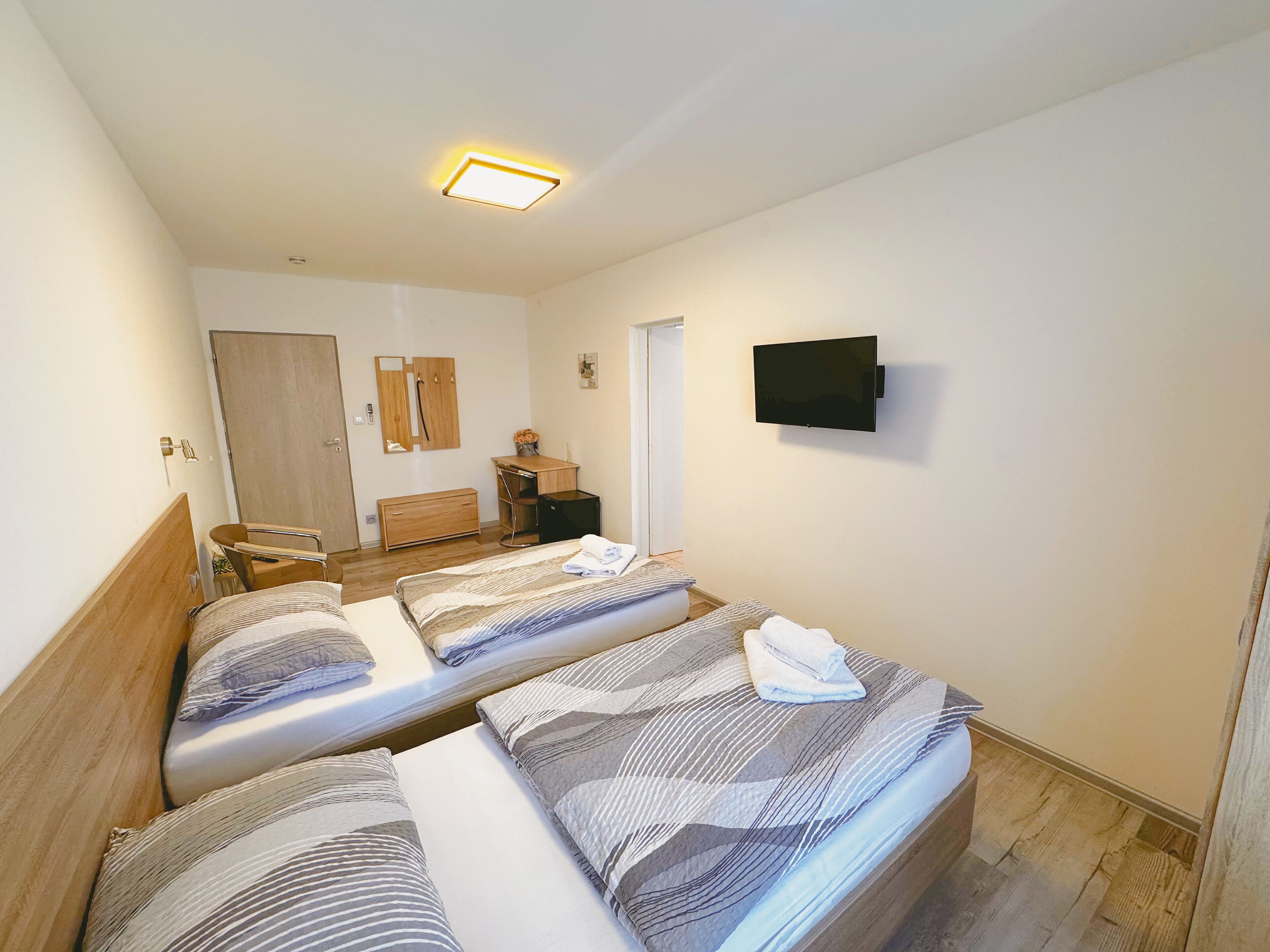 A hotel room with two beds, a wooden wardrobe, a desk with a chair, a television mounted on the wall, and a door leading to another room.
