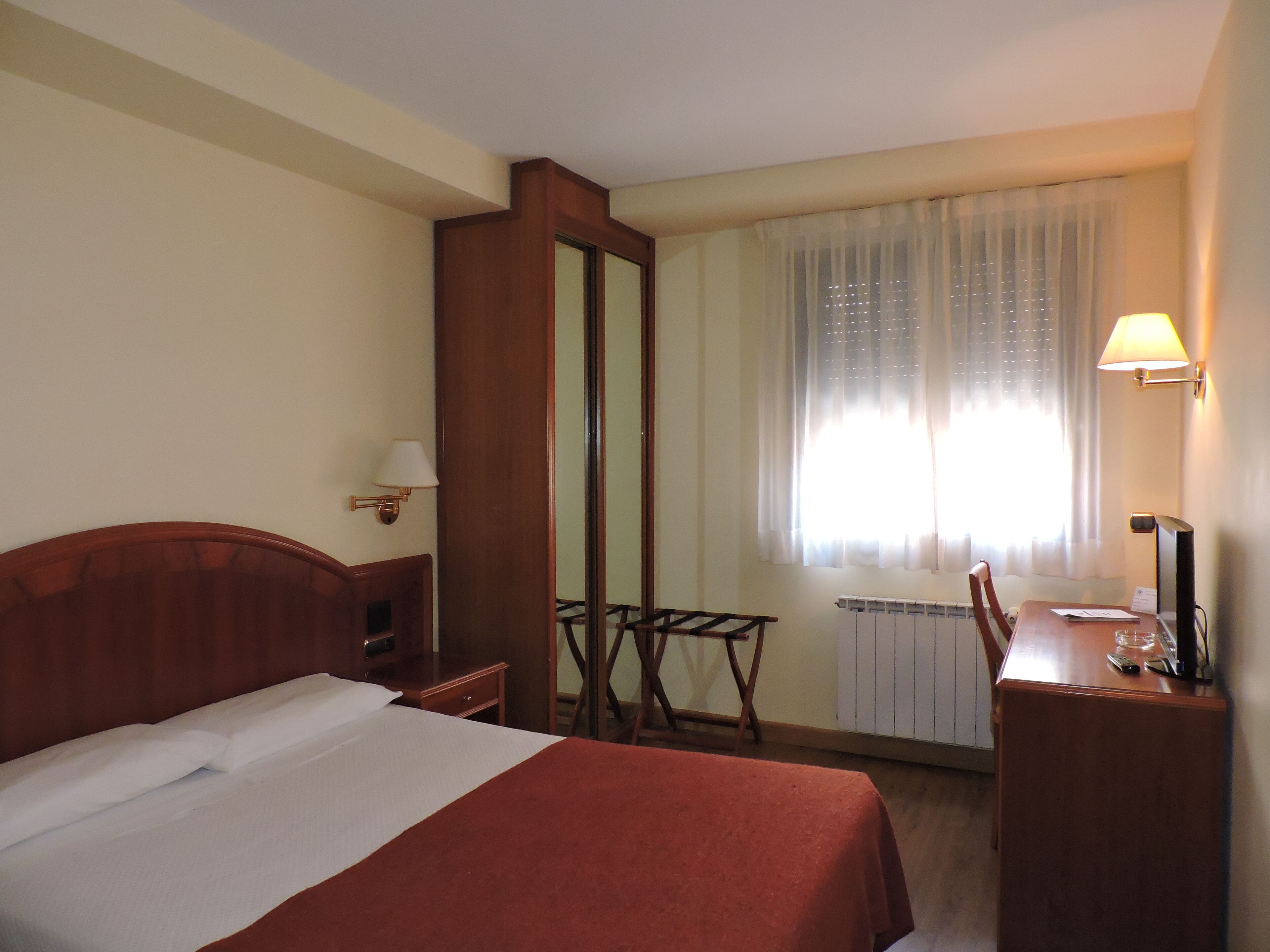 Habitación de hotel con cama, escritorio, silla, espejo y ventana con cortinas.