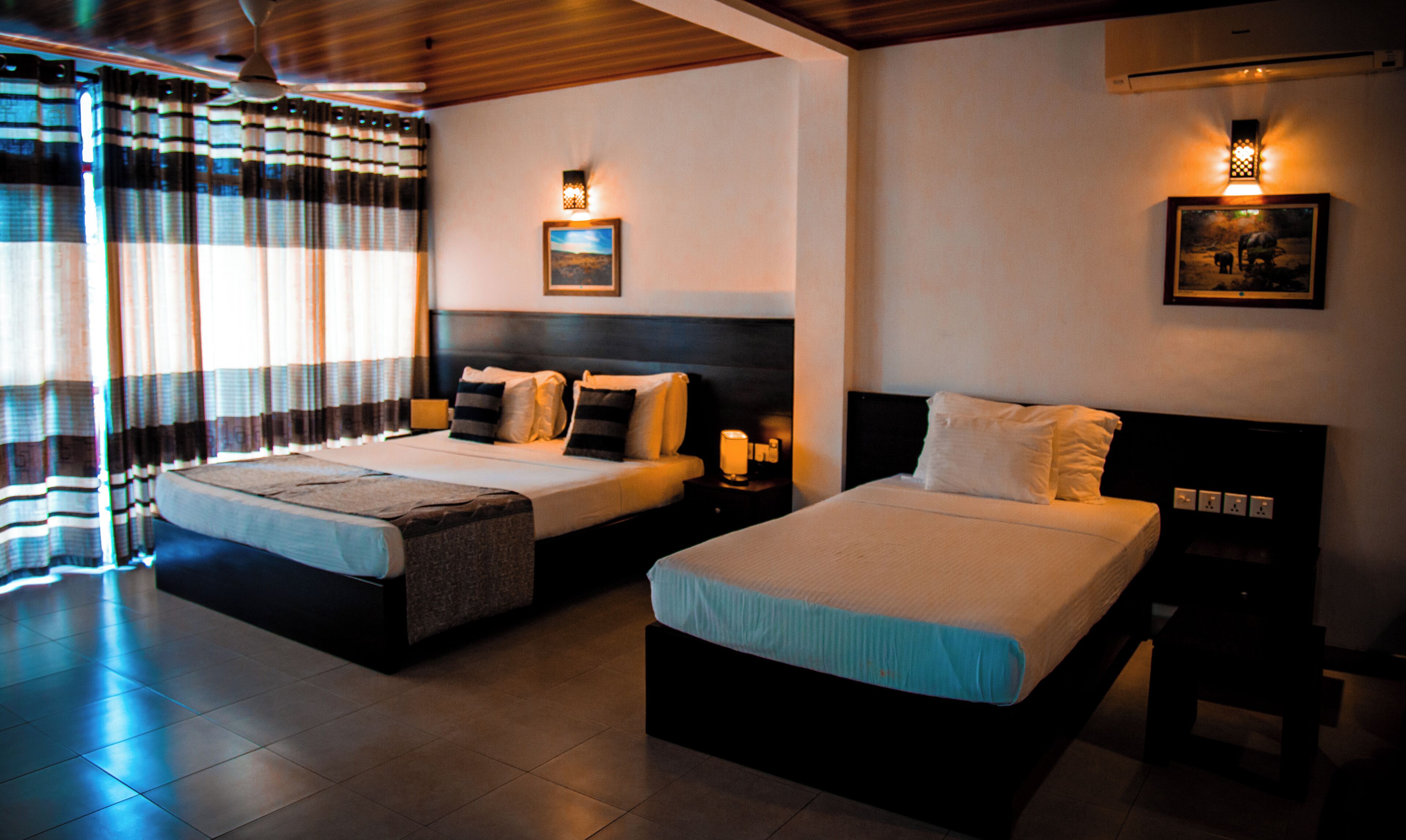 A hotel room with two beds, a large window with curtains, a painting on the wall, and a ceiling fan.