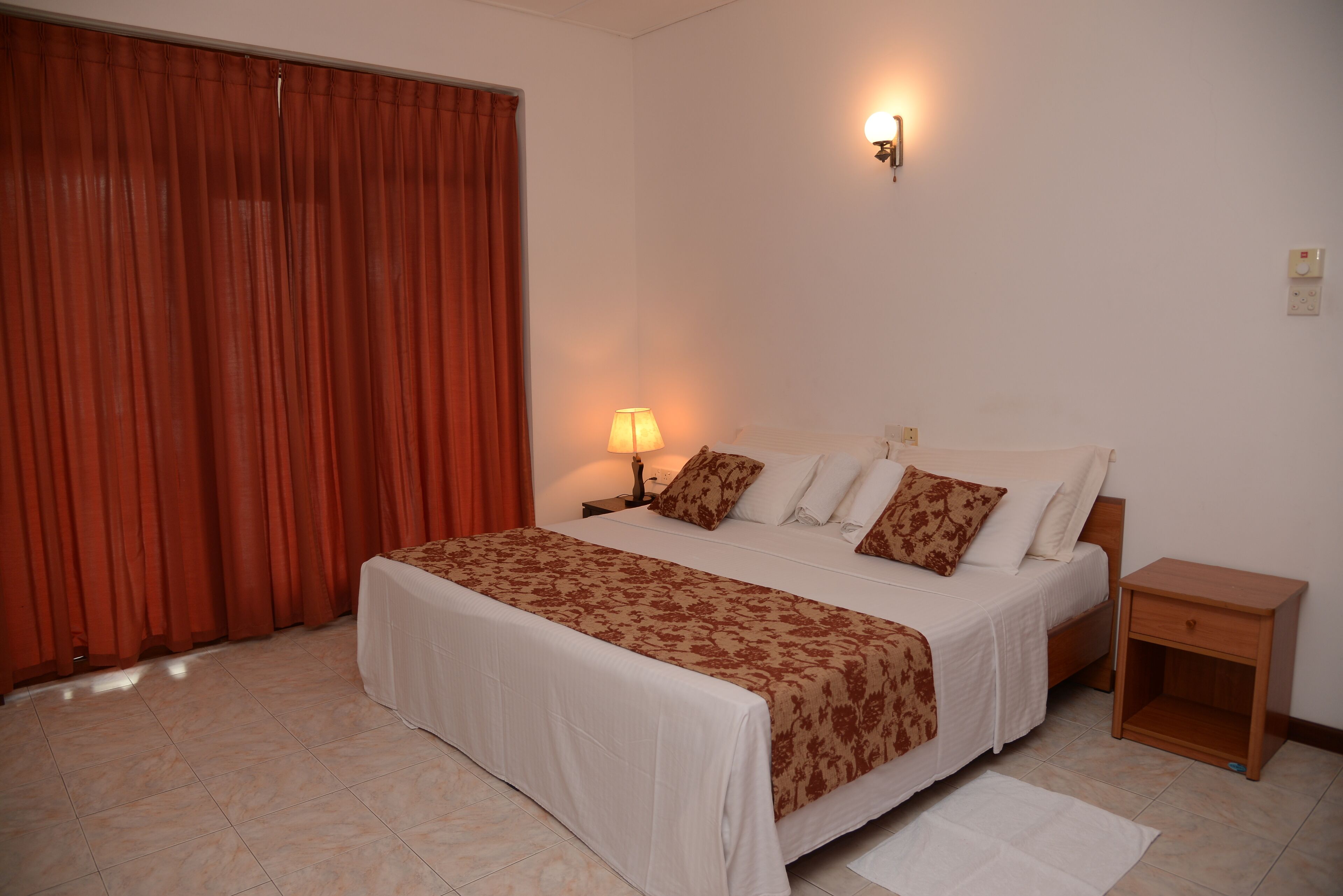 A bedroom with a bed, a nightstand, a lamp, a wall-mounted light, and a window with curtains.