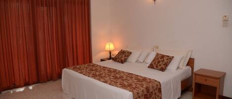 A bedroom with a bed, a nightstand, a lamp, a wall-mounted light, and a window with curtains.