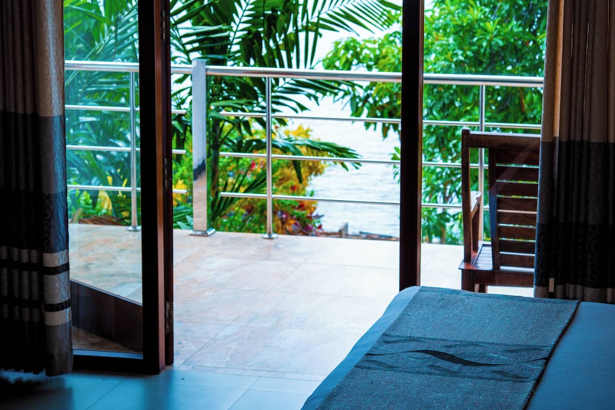 A room with a large sliding glass door leading to a balcony with a view of a body of water and greenery.