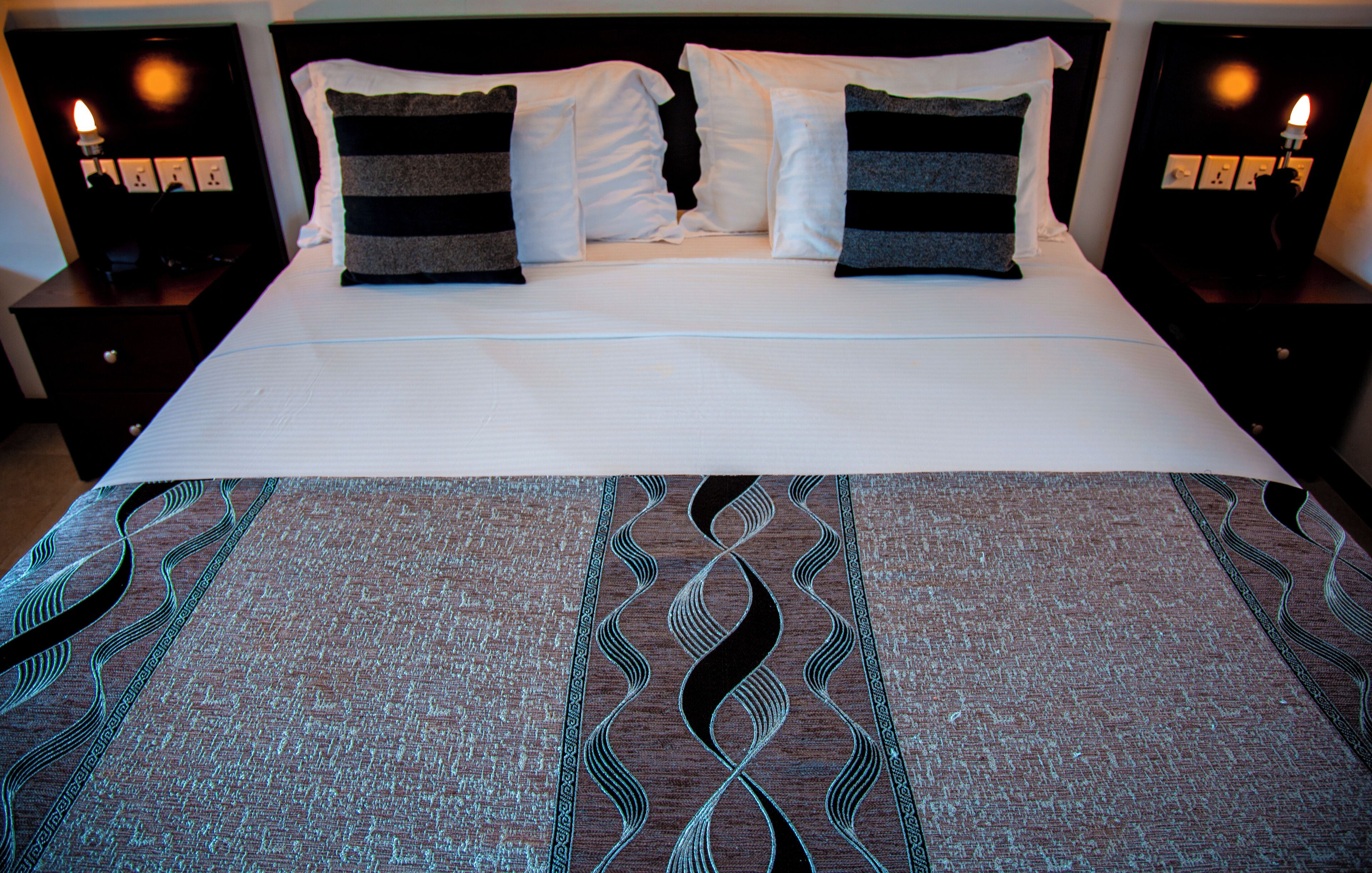 Un letto rifatto con cura, una coperta a motivi decorativi, due comodini con lampade e una testiera scura.