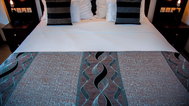 A neatly made bed with a patterned bedspread, two bedside tables with lamps, and a dark headboard. - Riverside Inn Fuji (Bentota)