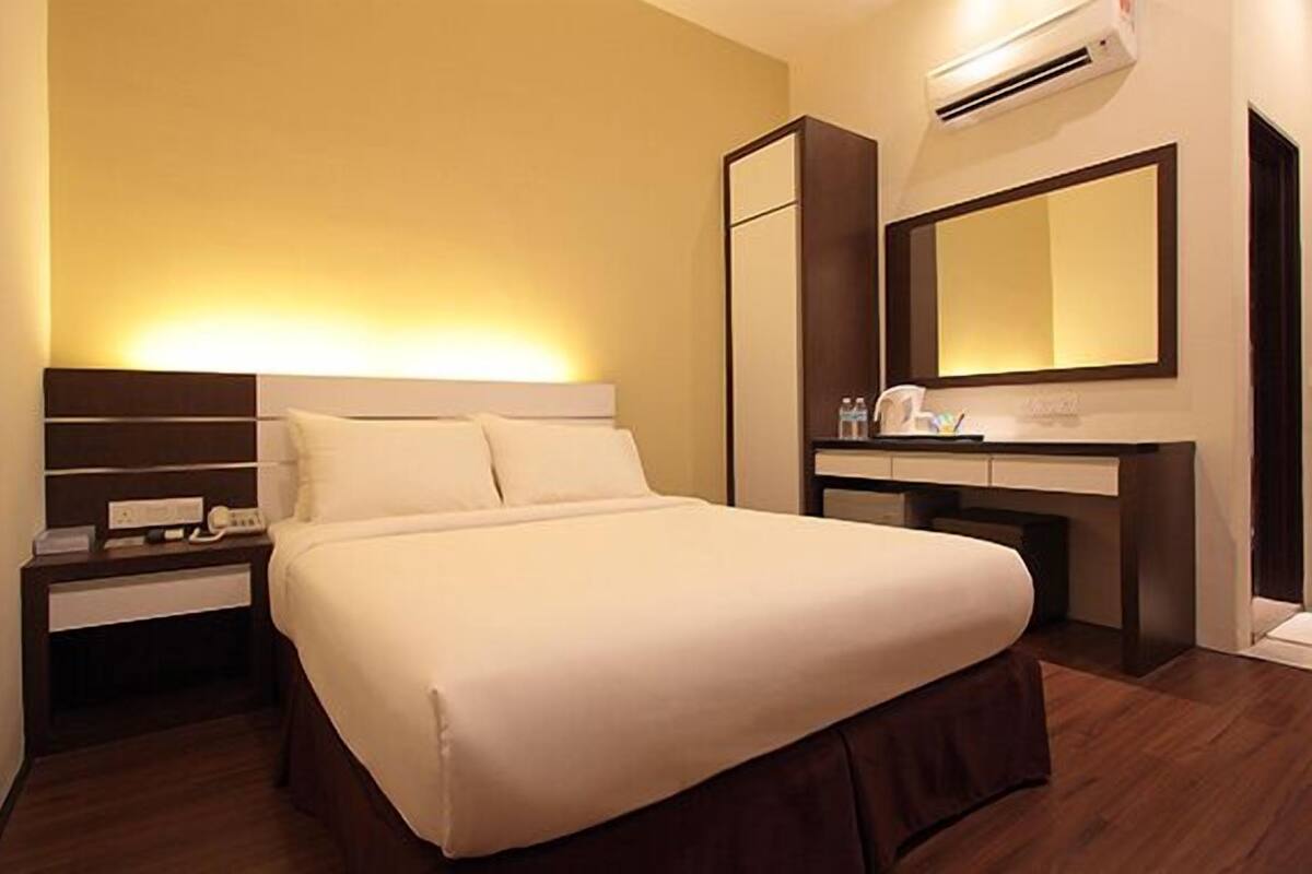 A hotel room with a bed, bedside table, lamp, mirror, and air conditioning unit.
