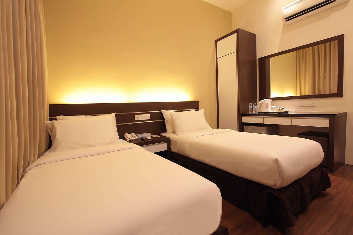 A hotel room with two beds, a bedside table with a phone and a lamp, a mirror, and an air conditioning unit.