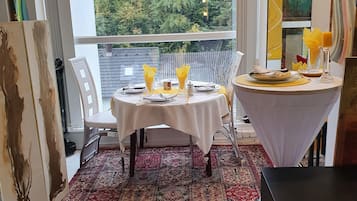 Un comedor con dos mesas dispuestas para dos comensales, cada una con mantel blanco, servilletas amarillas y velas. La habitación tiene un ventanal con vistas a árboles y un balcón.