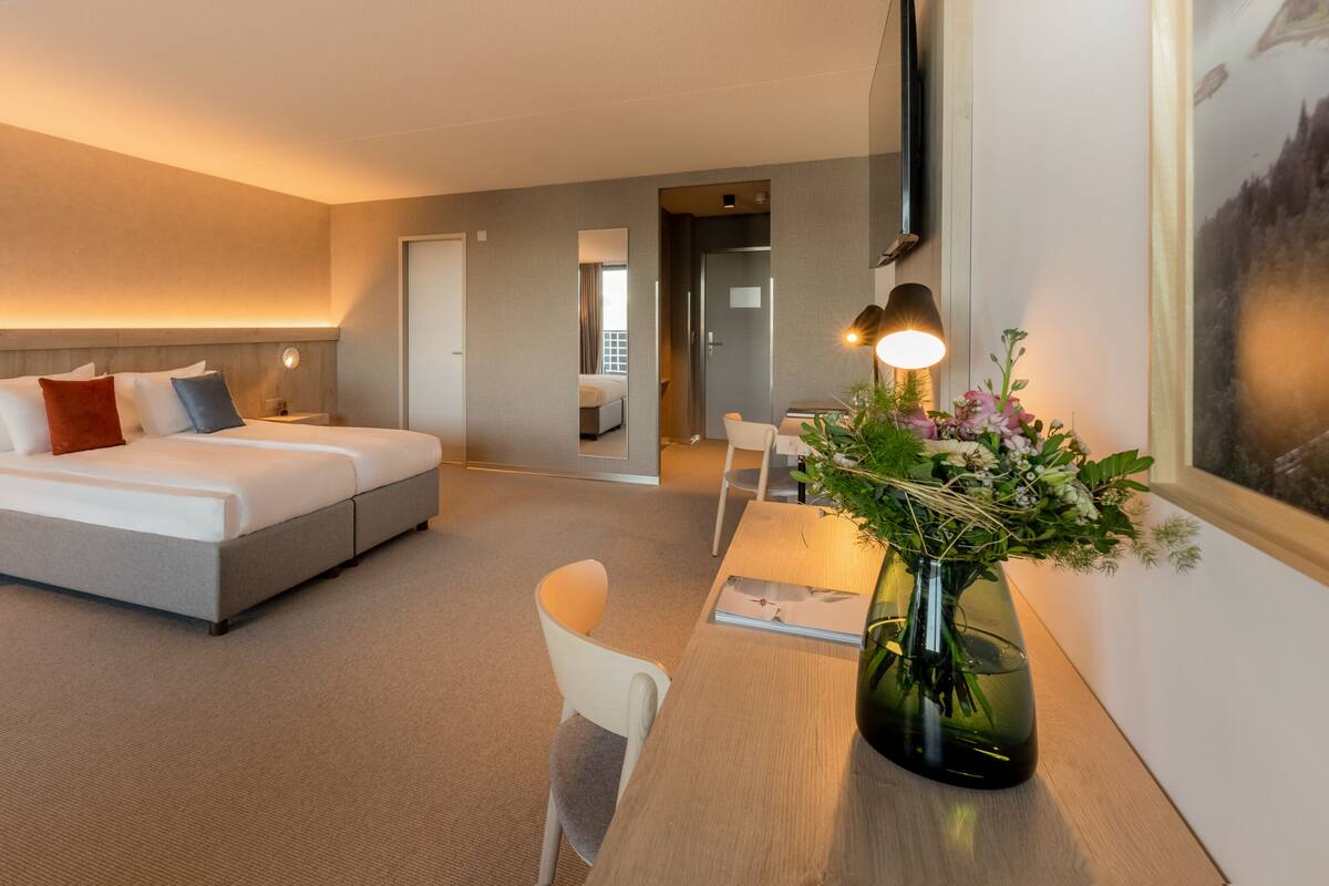 A modern hotel room with a large bed, a desk with a vase of flowers, and a view of the city through the window.