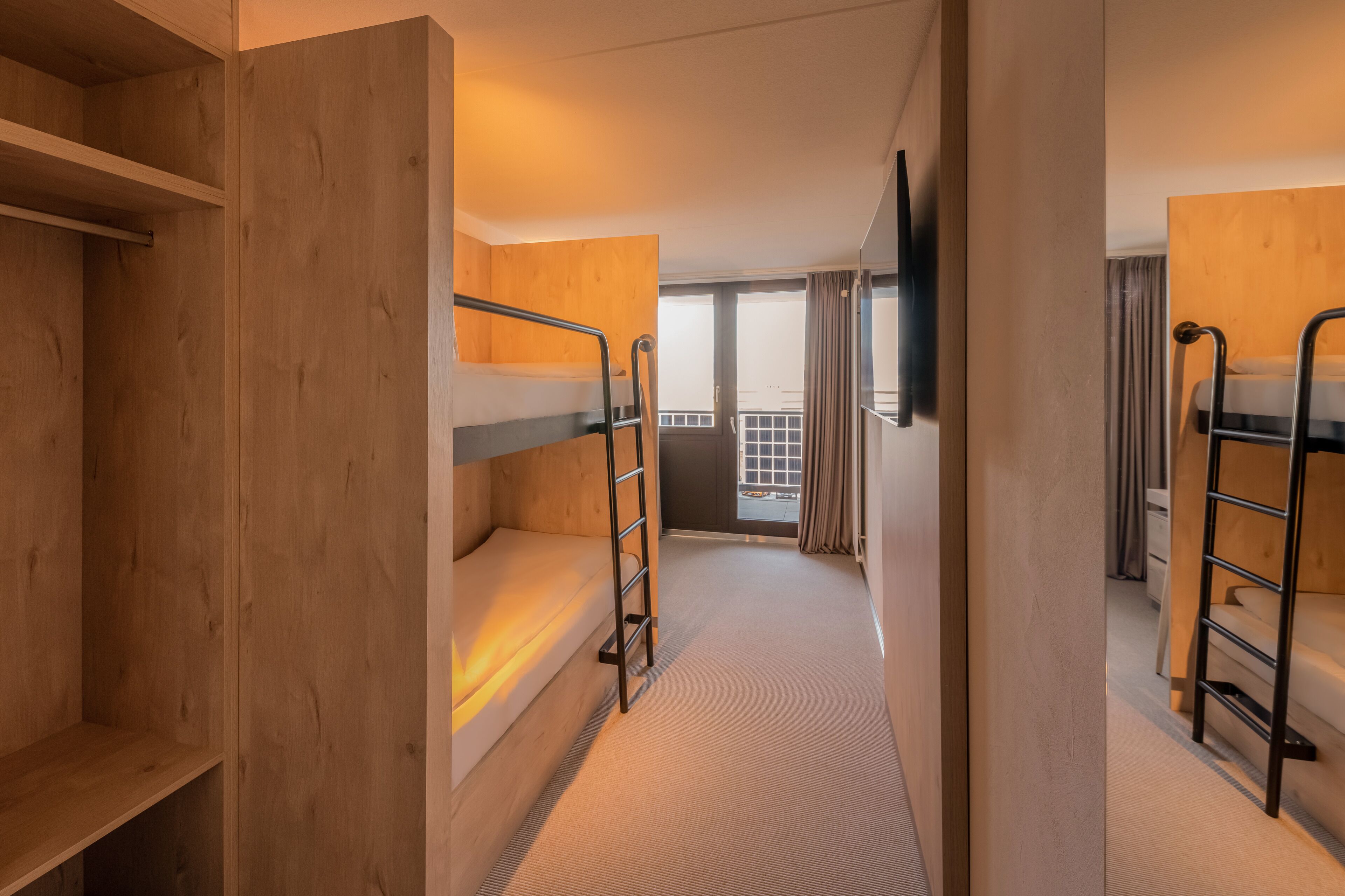 A hotel room with bunk beds, a wooden wardrobe, a television, and a balcony with curtains.