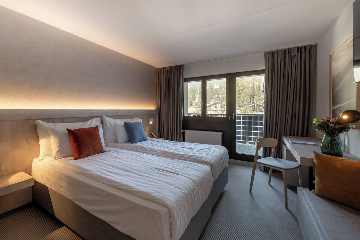 A hotel room with a large bed, a desk, and a chair. There is a window with curtains, and a view of trees outside.