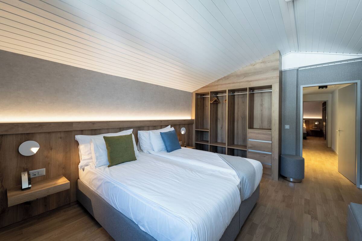 A modern hotel room with a large bed, wooden headboard, bedside tables with lamps, a wardrobe, and a walk-in closet.