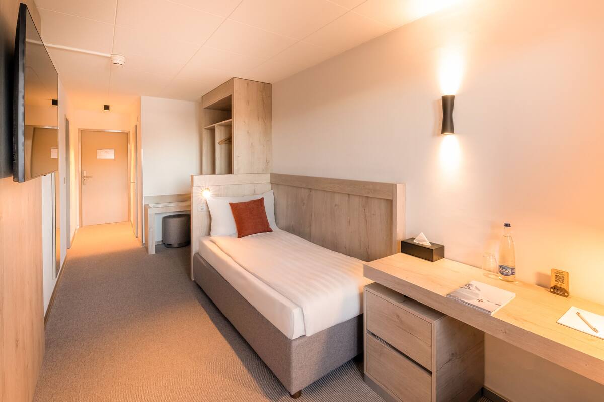 A hotel room with a bed, bedside table, and a desk with a chair. There is a TV mounted on the wall and a wooden headboard.