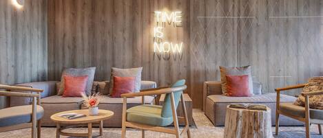 Ein modernes Wohnzimmer mit einer Holzvertäfelung, einem leuchtenden Schild mit der Aufschrift 'TIME IS NOW' und einer Mischung aus Sofas und Sesseln.