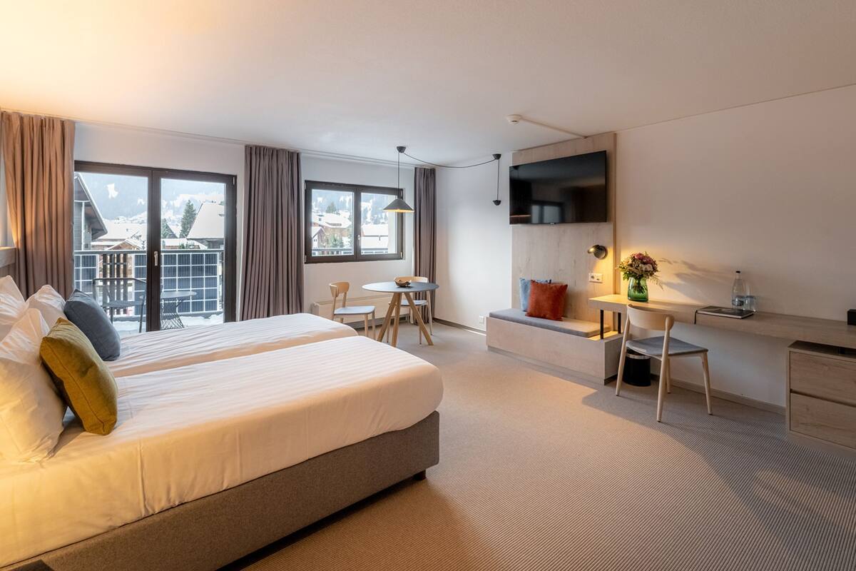 A hotel room with a bed, a desk, a chair, a TV, and a balcony with a view of the mountains.