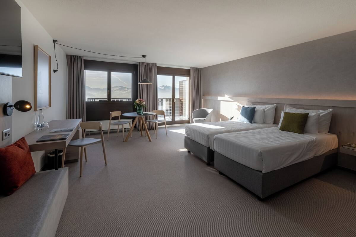 A modern hotel room with a large bed, a desk, and a chair. There is a balcony with a view of mountains.