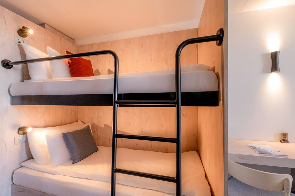 A bunk bed with a ladder, a desk, and a chair in a room with wooden walls and a white ceiling.