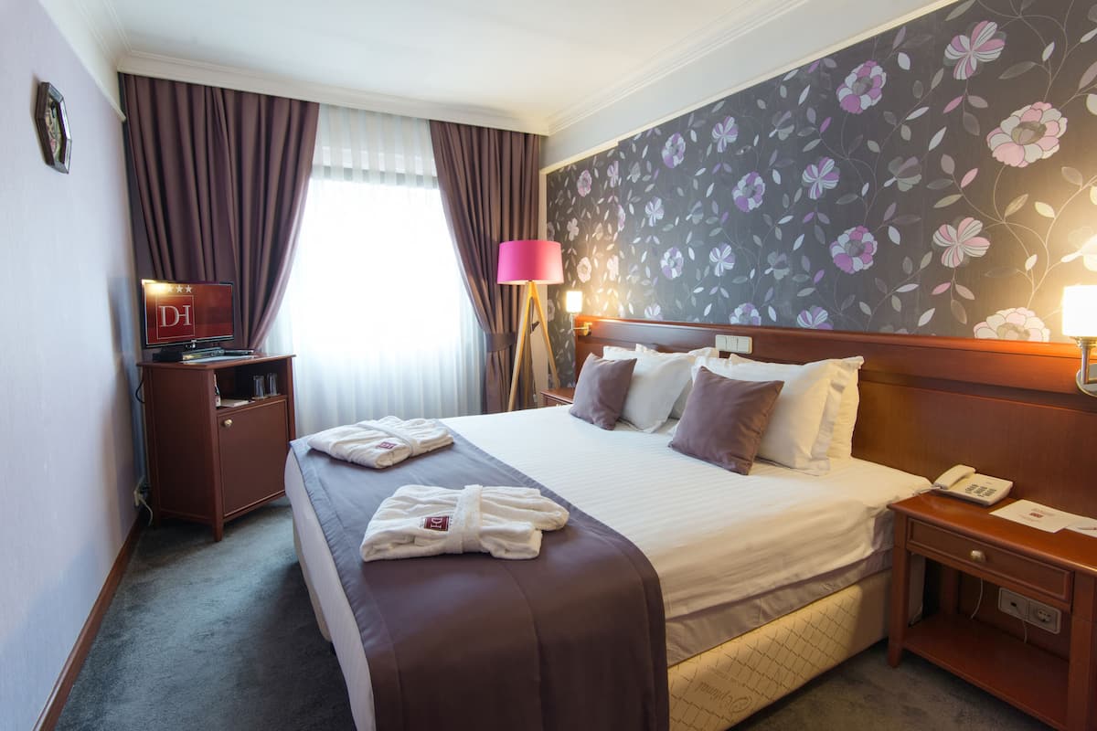 A hotel room with a bed, a bedside table with a lamp, a nightstand, a wardrobe, and a window with curtains.