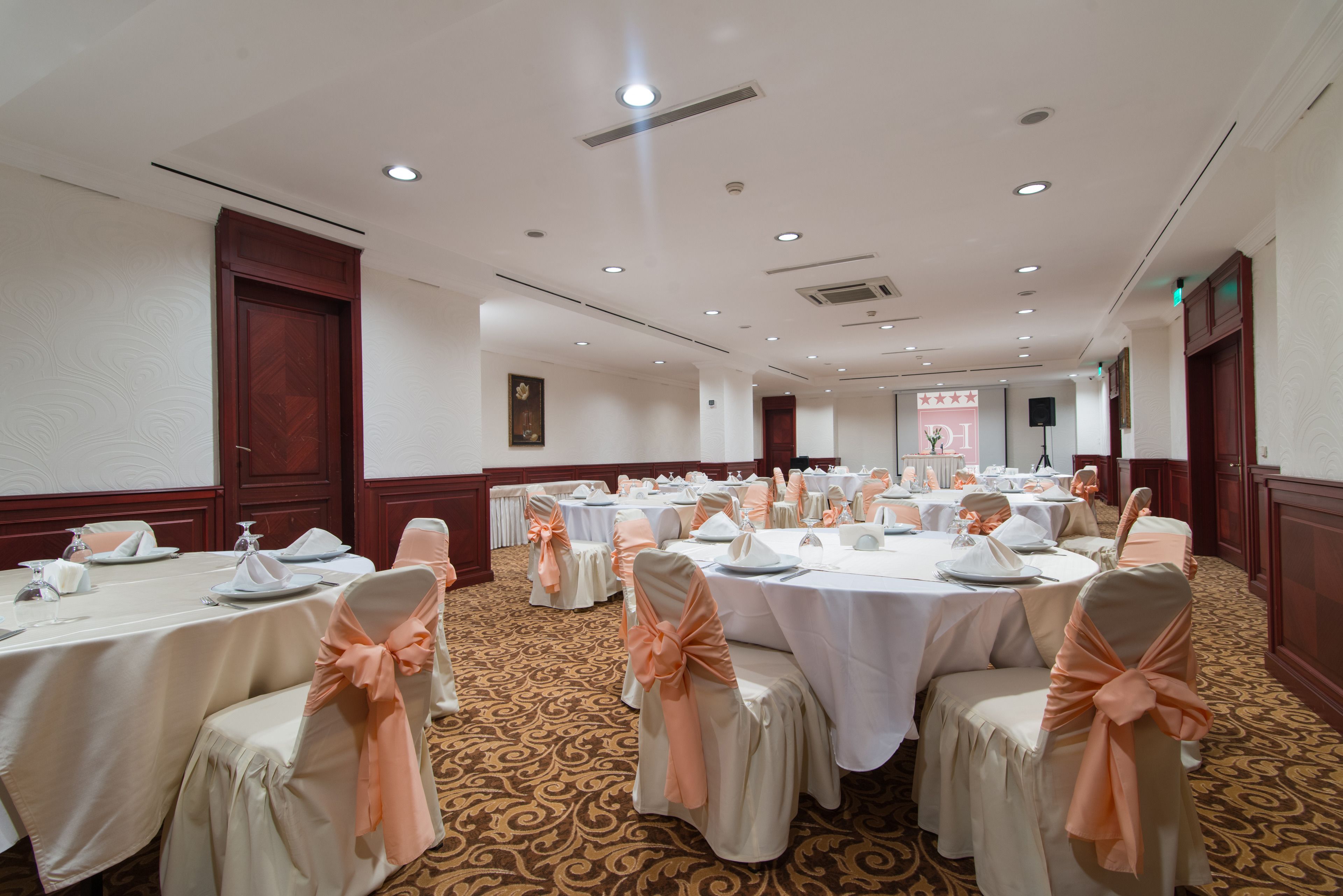 A banquet hall with round tables, chairs with peach-colored sashes, and a carpeted floor.