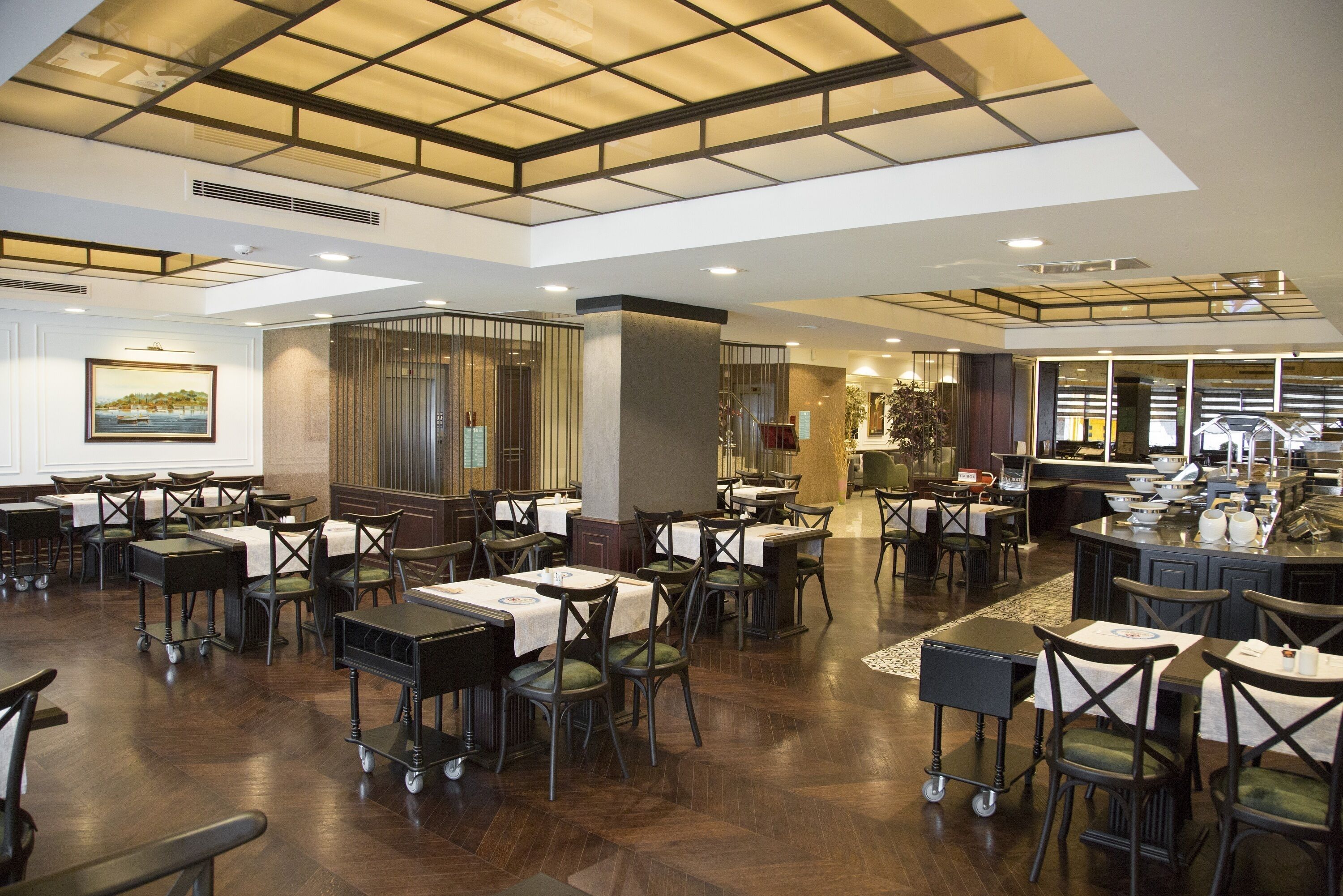 A spacious dining area with multiple tables and chairs, a high ceiling with skylights, and a kitchen in the background.