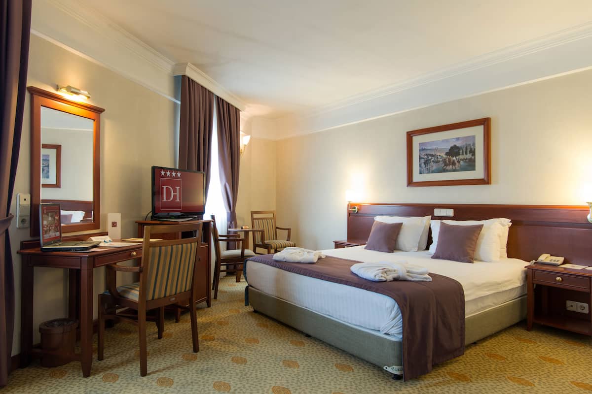 A hotel room with a bed, desk, chair, and a desk lamp.
