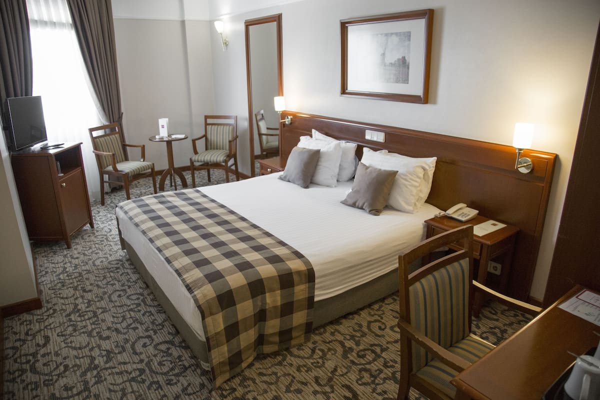 A hotel room with a large bed, a desk with a TV, two chairs, a small table, and a bedside lamp.