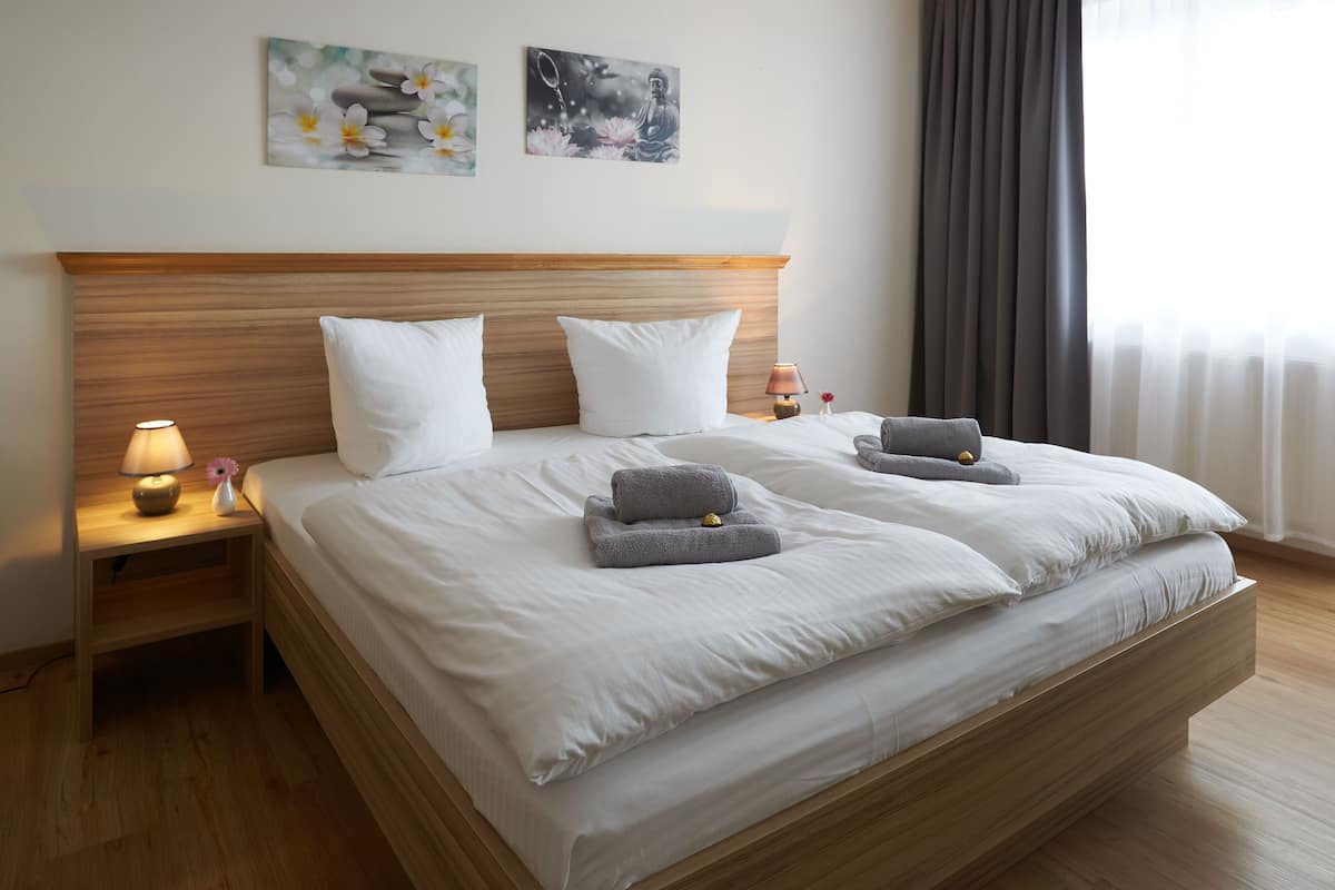 Una cama bien hecha con sábanas blancas, dos almohadas y dos toallas dobladas. Hay una mesita de noche con una lámpara y un florero con una flor. La habitación tiene cabecera de madera y un ventanal con cortinas.