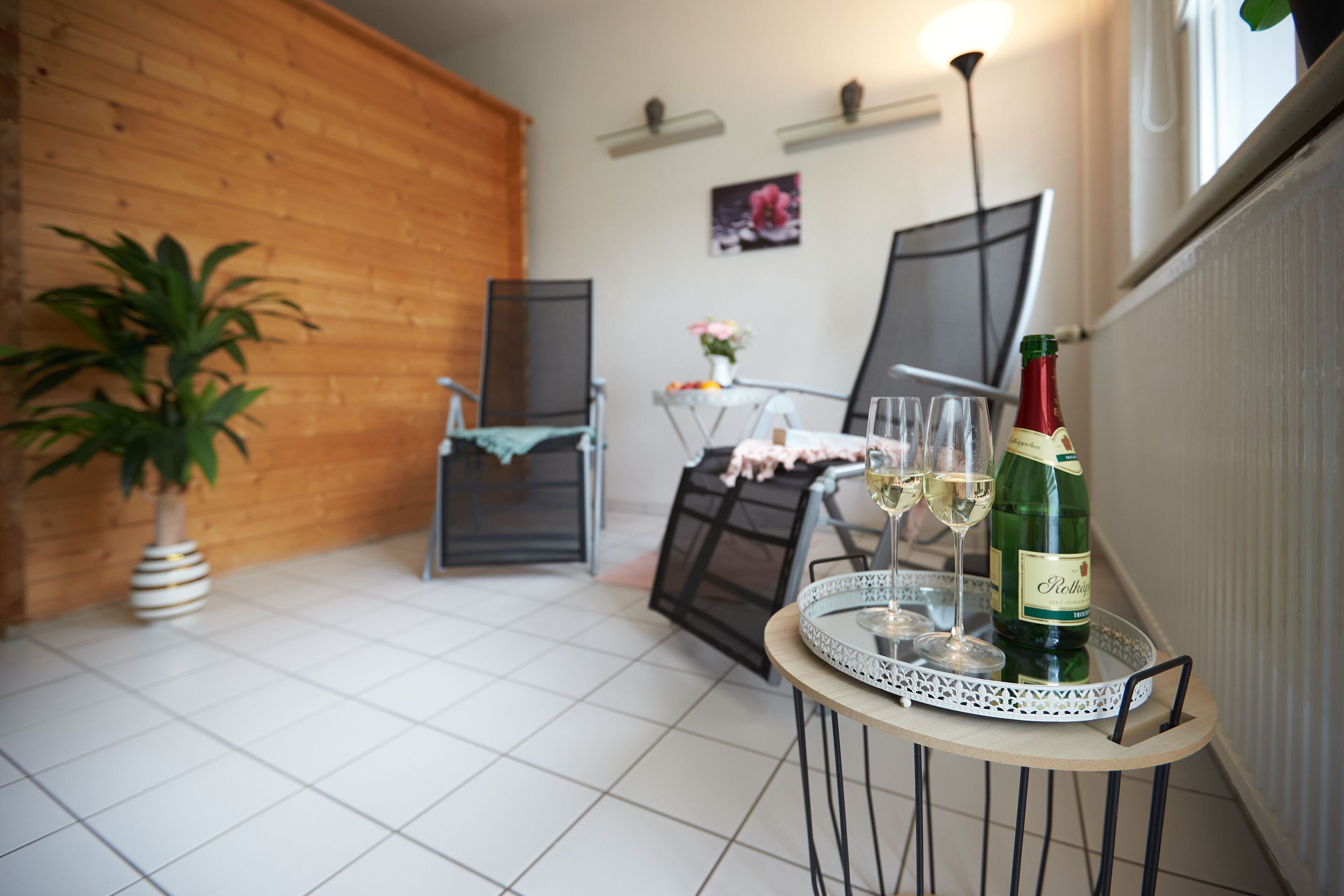 Une petite pièce accueillante, avec un sol carrelé, un mur en bois, une table surmontée d’un plateau en verre, une bouteille de vin et une chaise.
