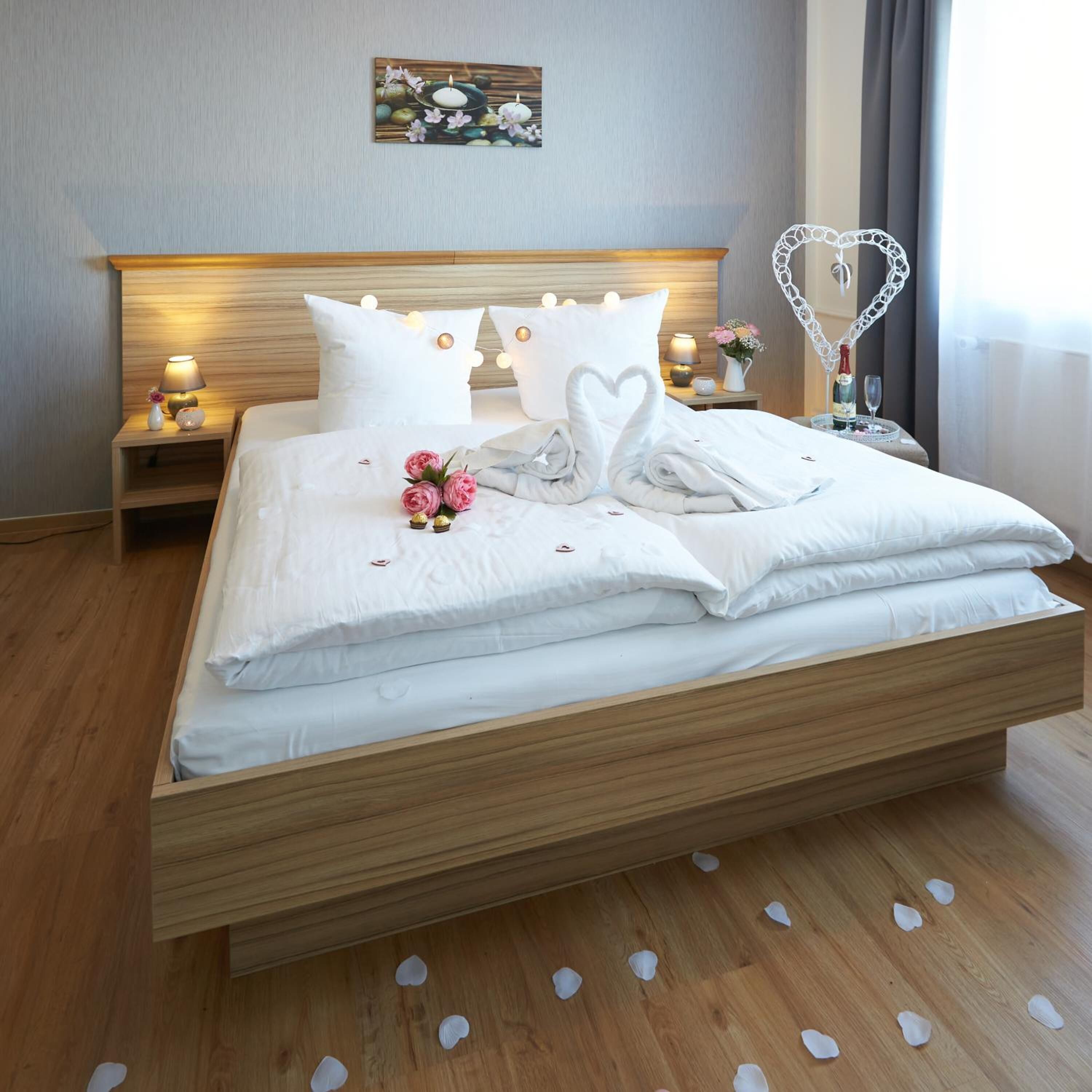 A neatly made bed with white linens, a heart-shaped pillow, and a bouquet of pink flowers.
