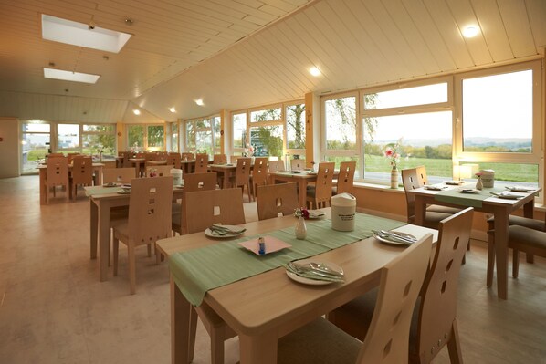 A spacious dining area with multiple tables and chairs, large windows offering a view of the outdoors, and a wooden ceiling. - Hotel Weimarer Berg (Apolda)