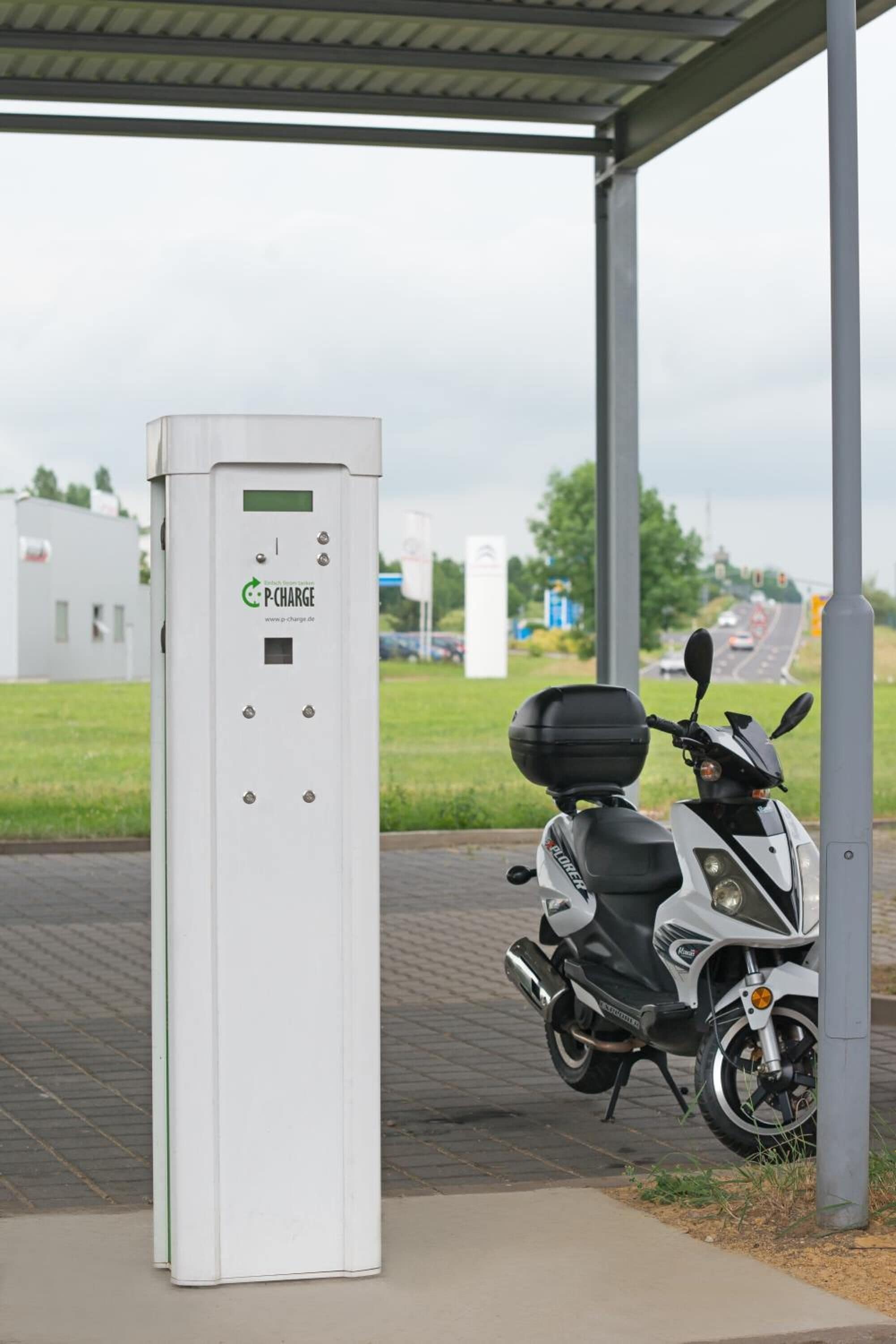 En P-charge el-scooterladestation med en hvid scooter parkeret i nærheden.