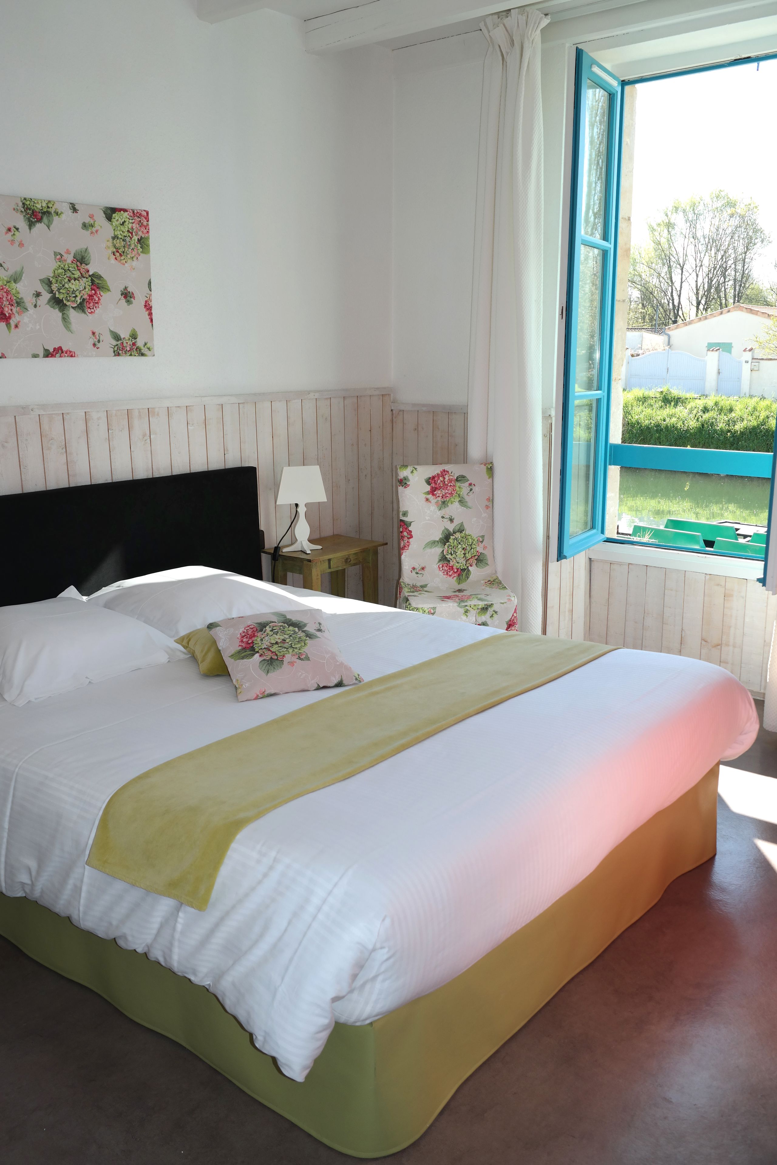 Un dormitorio con cama, mesita de noche, una silla, ventana con vista a follaje y una pared decorada con una pintura floral.