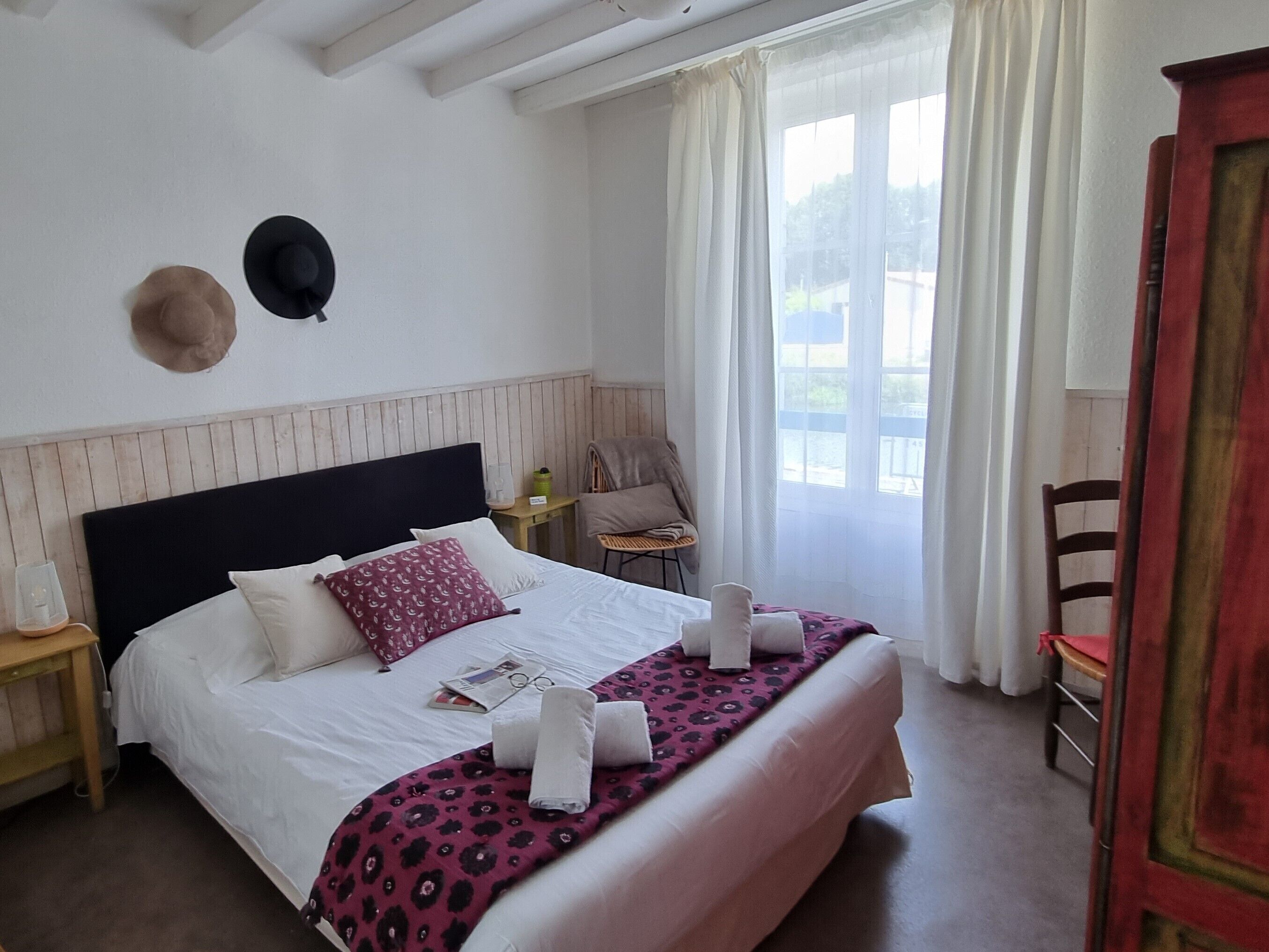Una cama bien hecha con sábanas blancas y una manta estampada, una mesita de noche con lámpara, y una ventana con cortinas de gasa.