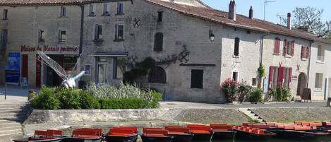 Un edificio histórico de piedra con techo de tejas, una fuente y varias embarcaciones amarradas en el agua de enfrente.